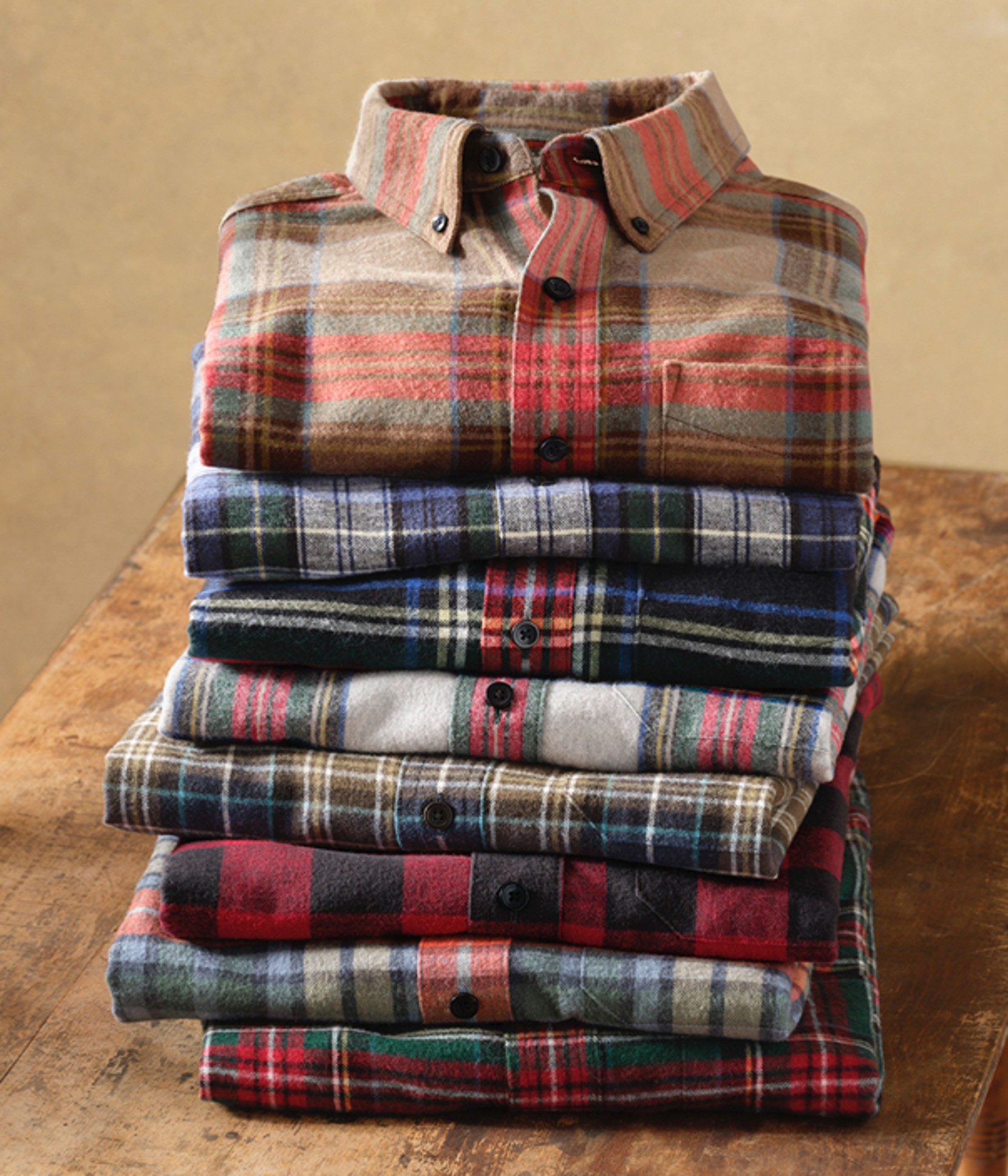 A stack of 8 folded plaid shirts in various colors, neatly arranged on a beige background.