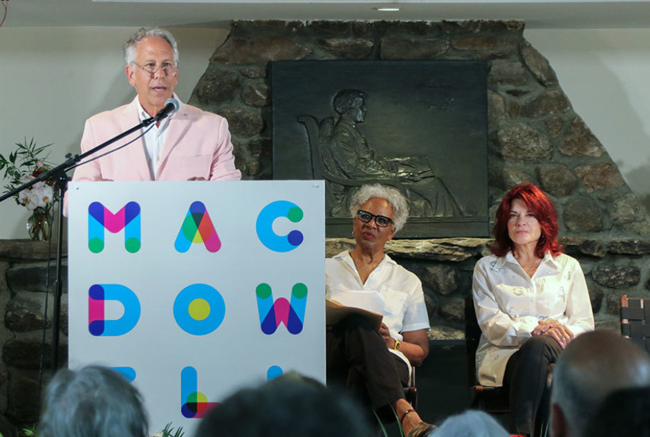 Phillip Himberg speaks at the podium on stage. Nell Painter and Roseanne Cash sit in chairs behind him.