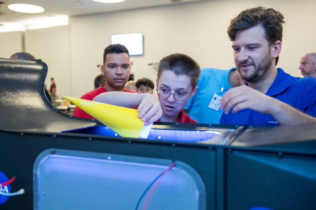 A researcher assists a student in guiding his cylindrical-shaped paper model into a tabletop demonstration unit.