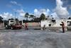 A flatbed truck with equipment on the back sits in a puddled parking lot near the remains of a demolished building foundation, with two figures in safety gear nearby and palm trees in the background. 