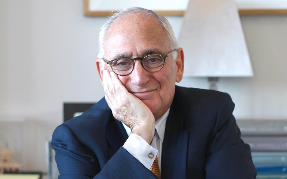 Robert A.M. Stern smiling and sitting on a desk with his hand on his chin