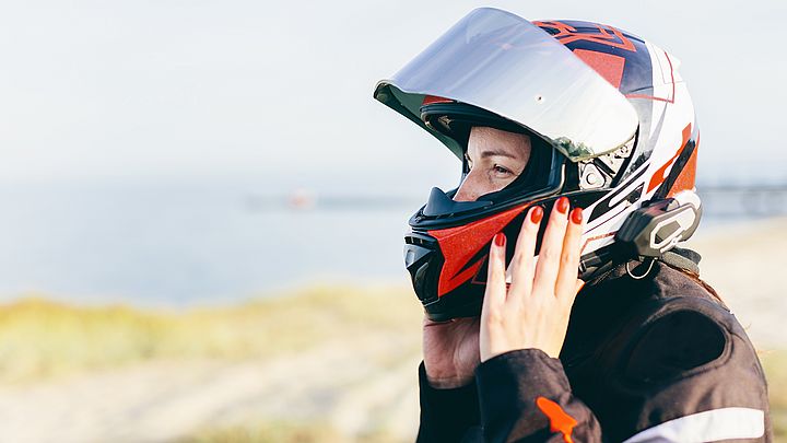 Eine Frau nimmt ihren Motorradhelm ab. Im Hintergrund ist dabei das Meer zu sehen