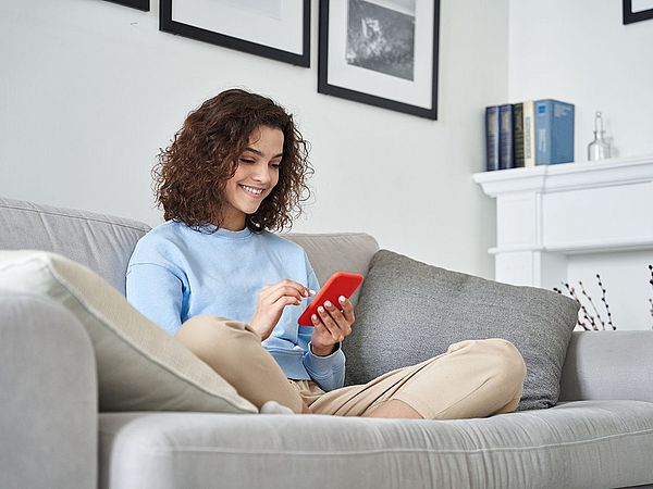 Eine Frau sitzt auf dem Sofa in ihrem Wohnzimmer und tippt dabei auf ihr Smartphone 