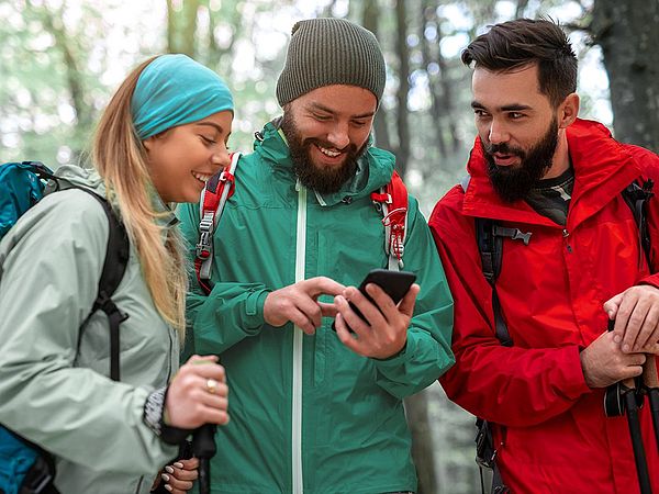 Eine Gruppe glücklicher junger Leute macht beim Wandern einen Halt, um den richtigen Weg zu suchen. Dabei wird das Smartphone als Unterstützung verwendet